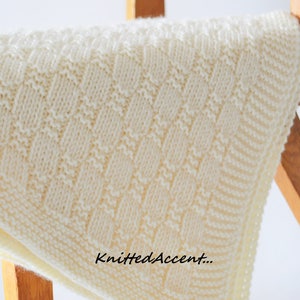 Puede incluir: Una manta de beb&eacute; de punto blanco con un patr&oacute;n de rombos. La manta est&aacute; doblada y drapeada sobre una silla de madera. El texto "KnittedAccent..." es visible en la esquina inferior derecha de la imagen.