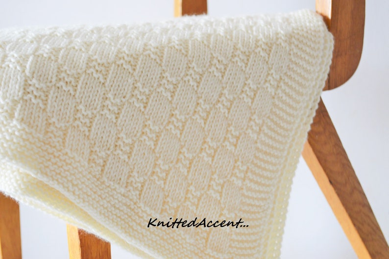 Puede incluir: Una manta de beb&eacute; de punto blanco con un patr&oacute;n de rombos. La manta est&aacute; doblada y drapeada sobre una silla de madera. El texto "KnittedAccent..." es visible en la esquina inferior derecha de la imagen.