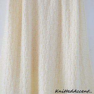 Puede incluir: Una manta de beb&eacute; de punto blanco con un delicado patr&oacute;n de puntadas. La manta est&aacute; colgada de una percha de madera y tiene el texto "Knitted Accent..." en la esquina inferior derecha.