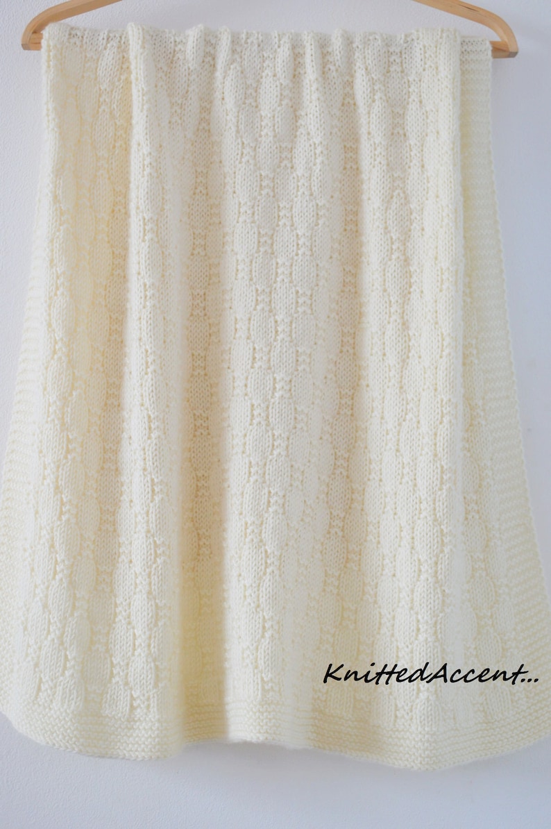 Puede incluir: Una manta de beb&eacute; de punto blanco con un delicado patr&oacute;n de puntadas. La manta est&aacute; colgada de una percha de madera y tiene el texto "Knitted Accent..." en la esquina inferior derecha.