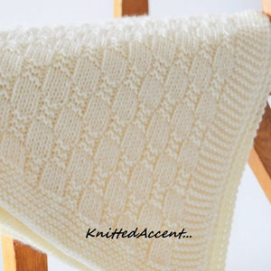 Puede incluir: Una manta de beb&eacute; de punto blanco con un patr&oacute;n texturizado. La manta est&aacute; drapeada sobre una silla de madera.