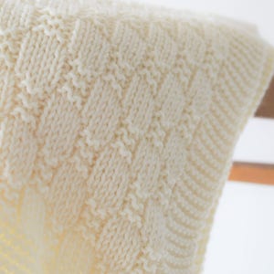 Puede incluir: Primer plano de una manta de punto blanca con un patr&oacute;n texturizado. La manta est&aacute; colgada en un estante de madera. El texto "Knitted Accent..." es visible en la esquina inferior derecha de la imagen.