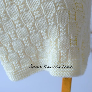 Puede incluir: Una manta de punto blanco con un patr&oacute;n de rombos. La manta est&aacute; doblada y drapeada sobre una silla de madera. El texto "Žana Daniūnienė..." es visible en la manta.