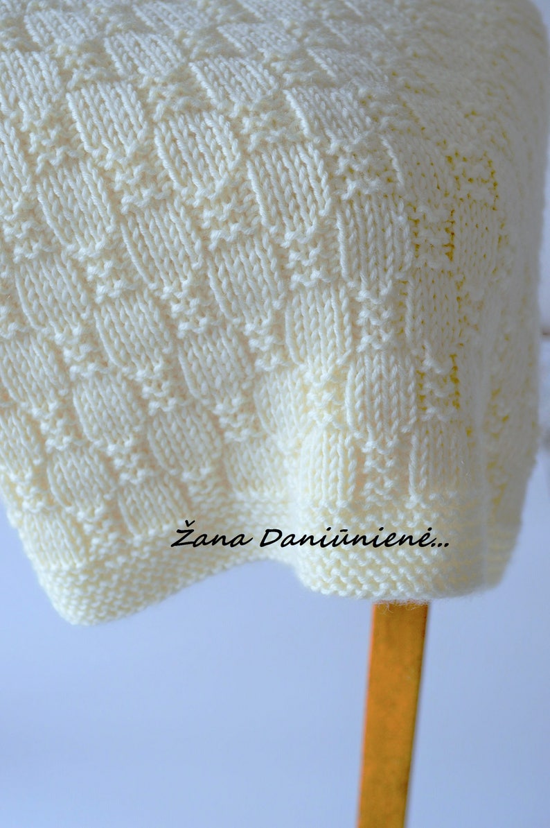 Puede incluir: Una manta de punto blanco con un patr&oacute;n de rombos. La manta est&aacute; doblada y drapeada sobre una silla de madera. El texto "Žana Daniūnienė..." es visible en la manta.