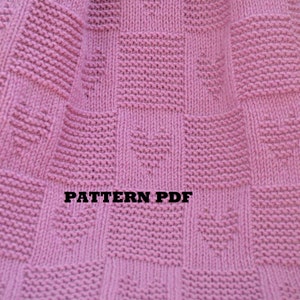 May include: A pink knitted blanket with a square pattern. The blanket has the text "PATTERN PDF" and "KnittedAccent" printed on it.