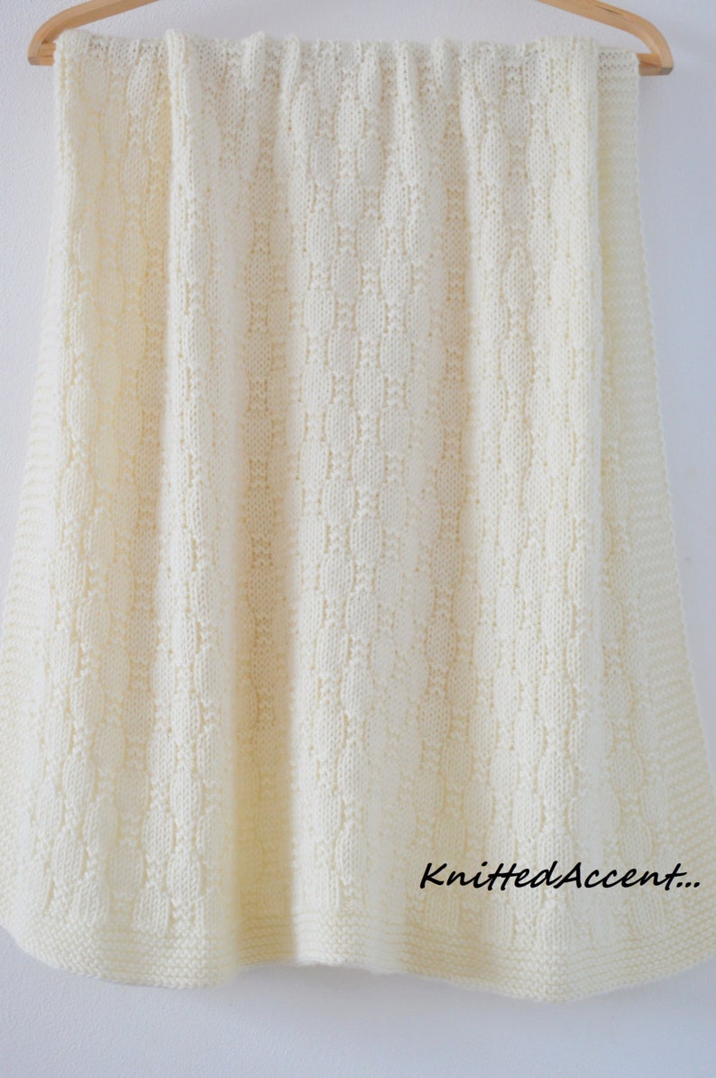 Puede incluir: Una manta de beb&eacute; de punto blanco con un delicado patr&oacute;n de puntada. La manta est&aacute; colgada de una percha de madera.