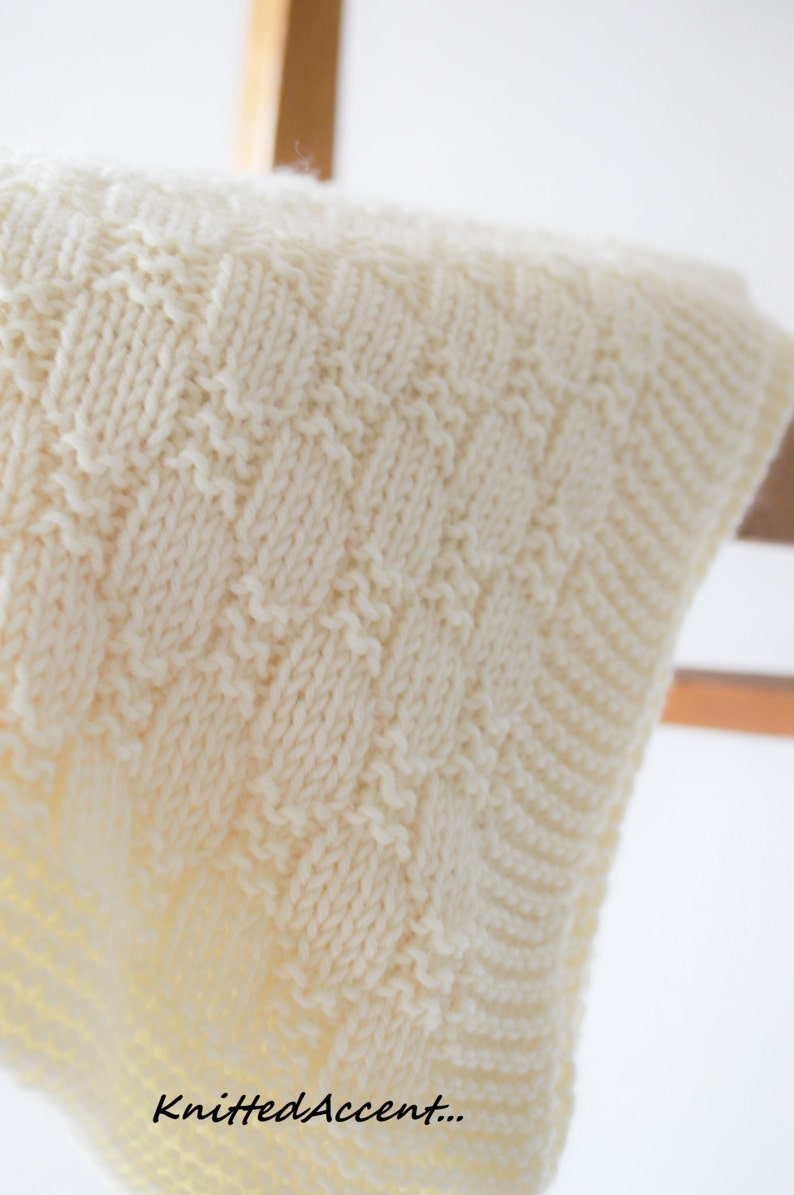 Puede incluir: Una manta de beb&eacute; de punto blanco con un patr&oacute;n texturizado. La manta est&aacute; colgada de una percha de madera. El texto "Knitted Accent..." es visible en la esquina inferior derecha de la imagen.
