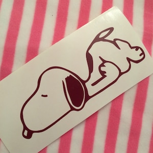 May include: A white sticker featuring a maroon outline of Snoopy lying down. The sticker is rectangular and has a glossy finish. The background is a pink and white striped pattern.