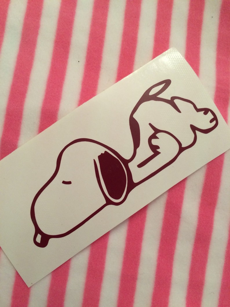 May include: A white sticker featuring a maroon outline of Snoopy lying down. The sticker is rectangular and has a glossy finish. The background is a pink and white striped pattern.