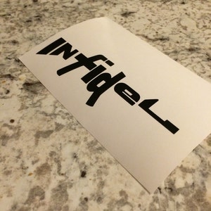 May include: A white sticker featuring the word "infidel" in a bold, black font. The sticker is angled, with the text in a stylized design. The sticker is placed on a speckled, light-coloured surface.