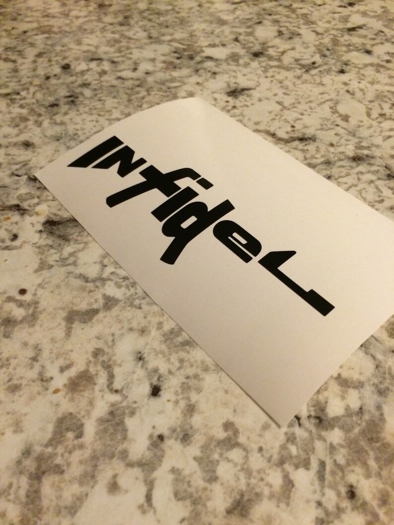 May include: A white sticker featuring the word "infidel" in a bold, black font. The sticker is angled, with the text in a stylized design. The sticker is placed on a speckled, light-coloured surface.