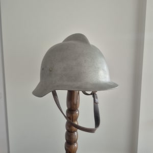 Könnte beinhalten: Ein metallisch silberfarbener Helm mit einem markanten Mittelkamm an der Oberseite, einer breiten Krempe und einem braunen Kinnriemen aus Leder, der an den Seiten befestigt ist. Der Helm hat eine glatte, gewölbte Form und eine matte Oberfläche.