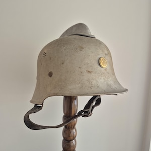 Könnte beinhalten: Ein vintage Metallhelm mit verwitterter grauer Oberfläche, ausgestattet mit einem markanten Mittelkamm, einem kleinen runden goldfarbenen Emblem an der Seite, Belüftungslöchern und einem dunkelbraunen Lederkinnriemen an der Helmschale.