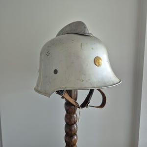 May include: A silver-coloured metal helmet featuring a prominent central ridge, a small circular gold emblem on the side, and a series of ventilation holes. The helmet includes a brown leather chin strap with metal buckles and hardware attachments.