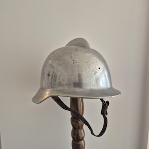 Può includere: Un elmetto color argento metallizzato con una prominente cresta centrale sulla parte superiore, una tesa larga e un sottogola in pelle marrone scuro con fibbia in metallo. La superficie dell'elmetto mostra segni di usura con piccoli graffi.