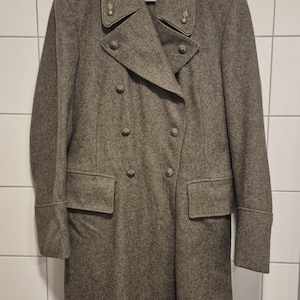 Op de afbeelding: Een lange, olijfgroene wollen jas met een dubbele rij knopen en decoratieve schouderstukken. De jas heeft twee rijen knopen, twee voorzakken en een revers. Hij hangt aan een witte hanger tegen een betegelde muur.