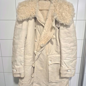 Puede incluir: Abrigo de invierno vintage color crema con cuello y forro de piel de oveja. El abrigo tiene cierre de botones, dos bolsillos delanteros y puños abotonados. El abrigo está colgado de una percha de madera.