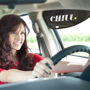 May include: A woman driving a car with a black sunshade that says "CHILL." The sunshade is attached to the windshield.