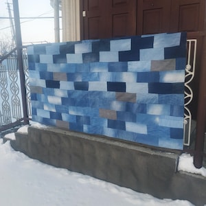 May include: A large rectangular denim patchwork quilt, displayed outdoors. The quilt is made of various shades of blue denim rectangles, creating a textured, geometric pattern. The quilt is hanging on a metal railing in front of a brown door.
