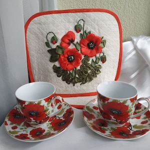 May include: Two white teacups and saucers with a red poppy floral pattern. A white tea cozy with red trim and a red poppy ribbon embroidery design.