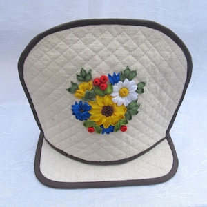 May include: A white quilted pot holder with a brown trim and a floral design embroidered with yellow, red, blue, and white ribbon.