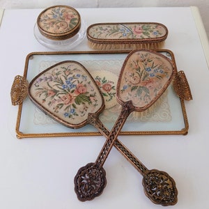Könnte beinhalten: Ein Set Vintage-Schminkutensilien, bestehend aus zwei Handspiegeln mit Blumenstickerei, einer Bürste mit Blumenmuster und einem kleinen Glasgefäß mit einem Blumendeckel.