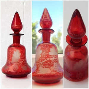 May include: Three red glass bottles with etched white grapevine designs. The bottles have a bell shape with a pointed top and a wide base. The bottles are all the same design but are different sizes.