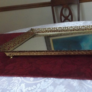 May include: A gold-colored, ornate, rectangular tray with a mirrored surface and decorative feet. The tray has a delicate, filigree design around the edge.