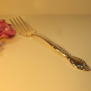 May include: A silver fork with an ornate design on the handle. The fork is lying on a white surface with pink flowers.