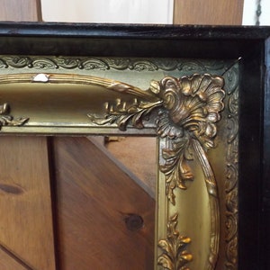 May include: A gold picture frame with ornate details and a black outer edge. The frame has a raised, curved top edge and a decorative floral design.