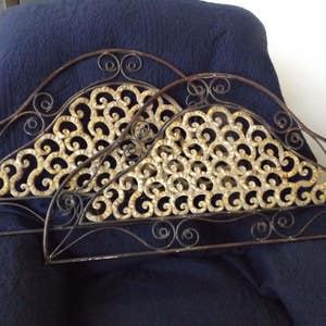 May include: Two decorative metal wall hangings in the shape of a fan. The fan is made of black metal with a gold-colored design. The fan is hanging on a blue fabric background.