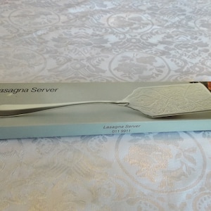May include: A silver lasagna server in its original box. The box is white with blue accents and the text "Lasagna Server" is printed on the front. The server has a decorative design and a long handle.