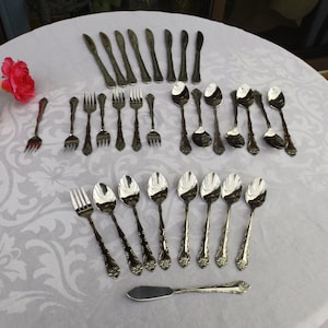 May include: A set of silverware laid out on a white tablecloth with a floral pattern. The set includes knives, forks, and spoons, all with ornate designs.