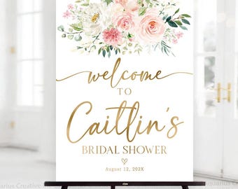 Puede incluir: Un letrero blanco con letras doradas y detalles florales, exhibido en un caballete negro. El letrero dice "Bienvenida a la despedida de soltera de Caitlin" con la fecha 12 de agosto de 202X. La parte superior presenta un arreglo floral de flores rosas y blancas.