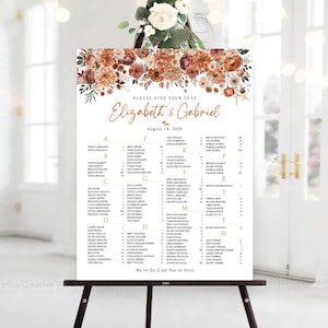 May include: A white wedding seating chart sign featuring a floral arrangement of rust, orange, and brown flowers at the top. The text reads Please Find Your Seat, Elizabeth & Gabriel, August 18, 20XX, and lists guest names organized by table letters.