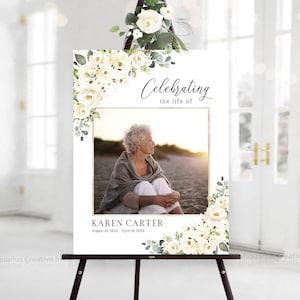 Könnte beinhalten: Ein weißes Schild auf einer schwarzen Staffelei, verziert mit weißen Blumengestecken. Das Schild zeigt ein Foto und den Text "Celebrating the life of Karen Carter". Das Schild ist mit weißen Rosen und Grün geschmückt.