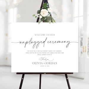 Puede incluir: Un letrero blanco sobre un caballete negro con el texto "Welcome to our unplugged ceremony." El letrero también incluye los nombres Olivia & Jordan y la fecha 18 de agosto de 2025. El letrero está decorado con flores blancas y vegetación.