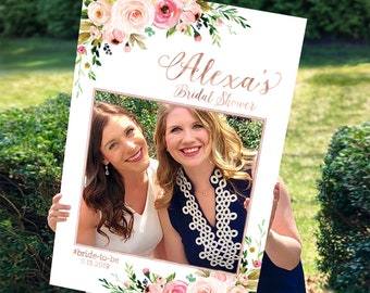 May include: A white photo booth prop with a floral design and a frame for a picture. The text "Alexa's Bridal Shower" is written in rose gold lettering. The hashtag "#bride-to-be" and the date "9.15.2019" are written in rose gold lettering at the bottom of the prop.