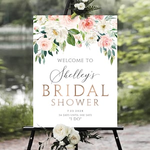 May include: A white sign with a floral border and gold lettering announcing a bridal shower for Shelley. The sign says "Welcome to Shelley's Bridal Shower 7.23.202X 54 Days Until She Says "I Do""