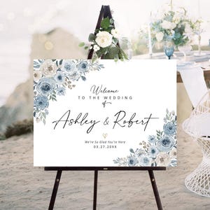 Peut inclure: Panneau de bienvenue de mariage blanc sur un chevalet noir, orn&eacute; de motifs floraux bleus. Le panneau indique "Bienvenue au mariage de Ashley & Robert" et la date 03.27.20XX. Un arrangement floral est plac&eacute; au-dessus du chevalet.