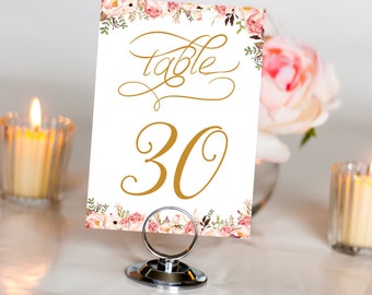 May include: A white table number card with a floral border and gold lettering. The card says "table" and "30". The card is on a silver stand.