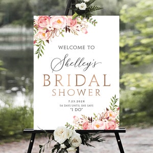 May include: A white bridal shower sign with pink and green floral decorations. The sign reads "Welcome to Shelley's Bridal Shower 7.23.202X 54 Days Until She Says "I Do""