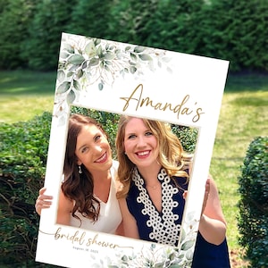 May include: A white photo booth prop with a gold frame and green foliage. The text "Amanda's bridal shower August 18, 2023" is printed on the prop.