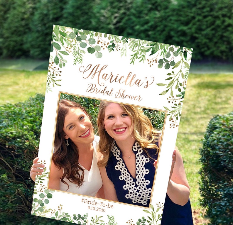 May include: A white photo booth prop with a gold frame and green floral border. The text "Mariella's Bridal Shower #Bride-To-Be 9.16.2019" is printed on the prop.
