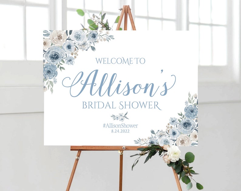 Op de afbeelding: Een wit welkomsbord met blauwe en witte bloemenmotieven. Het bord luidt "Welkom bij Allison's Bridal Shower #AllisonShower 8.24.2022".