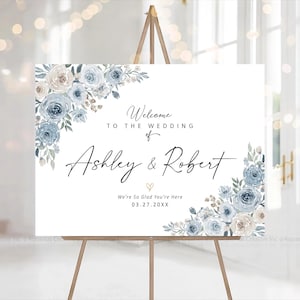 Peut inclure: Panneau de bienvenue de mariage blanc sur chevalet en bois. Il pr&eacute;sente des arrangements floraux bleus et cr&egrave;me. Le texte indique &laquo;&nbsp;Bienvenue au mariage de Ashley & Robert&nbsp;&raquo; et la date de l'&eacute;v&eacute;nement.