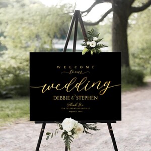 Puede incluir: Un letrero de bienvenida de boda negro con letras doradas sobre un caballete de madera. El letrero dice "WELCOME to our wedding DEBBIE & STEPHEN Thank You FOR CELEBRATING WITH US August 17, 202X". Flores blancas y vegetaci&oacute;n adornan la parte superior e inferior.