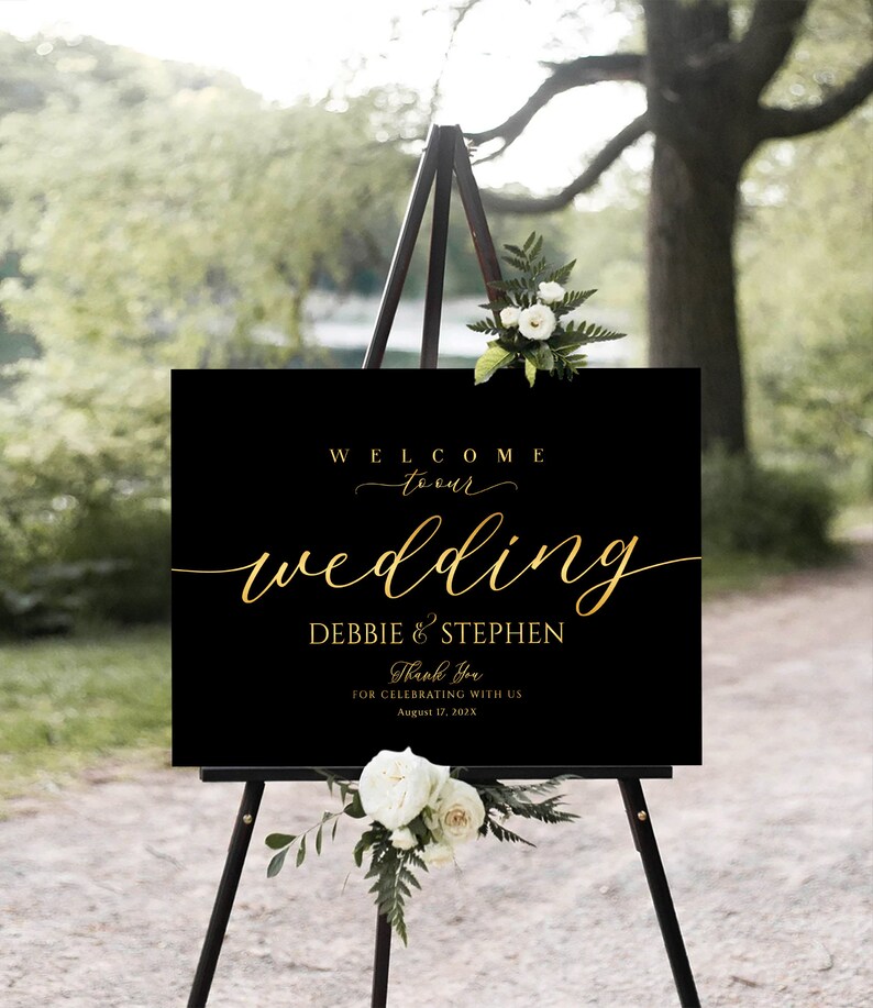 Puede incluir: Un letrero de bienvenida de boda negro con letras doradas sobre un caballete de madera. El letrero dice "WELCOME to our wedding DEBBIE & STEPHEN Thank You FOR CELEBRATING WITH US August 17, 202X". Flores blancas y vegetaci&oacute;n adornan la parte superior e inferior.