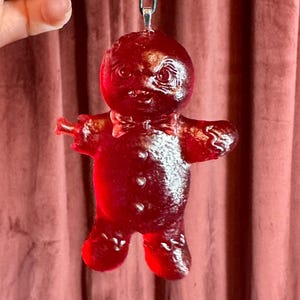 May include: A translucent red gingerbread man ornament with a bow tie and outstretched arms. The ornament is suspended by a thin green string against a pinkish-red curtain backdrop. The gingerbread man has a glossy finish.