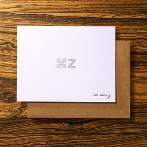 May include: A white card with a Command symbol and the letter "Z" in gray, with the words "I'm sorry" in black script. The card rests on a brown envelope, set against a wooden background.
